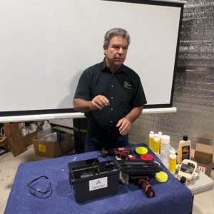 Mike Ello presenting beside a projection screen, with a UV curing lamp case, polish bottles and a cordless polisher set out on the table.